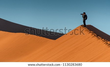 Young male traveler and photographer standing on the top of sand dune photographing sunrise or sunset in desert of Namibia, Africa. Travel photography concept