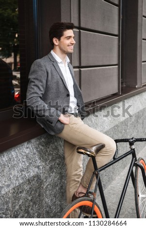 Cheerful young businessman leaning on a wall while standing outdoors with a bicycle