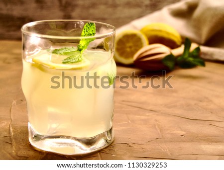 25 degrees picture of glass of lemonade with lemon, ice cubes and peppermint inside, and lemons, peppermint and wood squeezer at the back, over brown surface and wood background