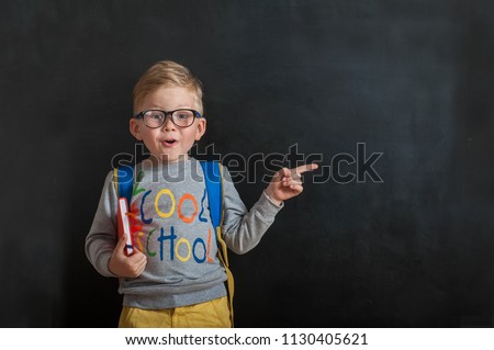 Back to school. Funny little boy in glasses pointing up on blackboard. Child from elementary school with book and bag. Education. Kid with a book