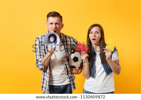 Expessive crazy couple, woman man football fans screaming, upset of loss, goal of favorite team with soccer ball, red card, megaphone isolated on yellow background. Sport family lifestyle concept