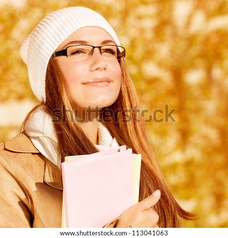 Picture of romantic student girl with textbook on yellow foliage background, closeup portrait of smart female wearing warm hat and stylish glasses, back to school, education concept