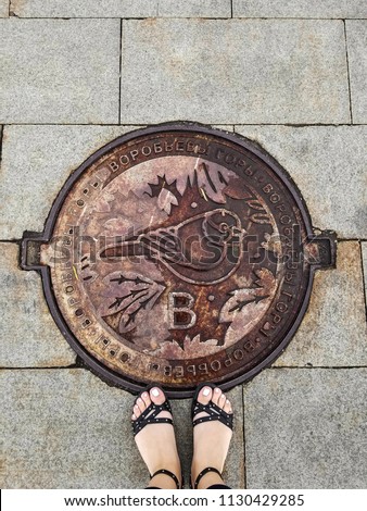 Girl's feet stand on sewer manhole on Vorobyovy Gory. Bird on hatch is a sparrow - symbol of place.