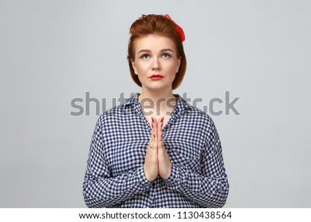Human emotions, feelings and reaction. Stylish young European female in checkered shirt looking up with mournful hopeful facial expression, keeping hands in namaste, praying. Please forgive me