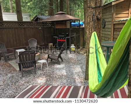 Hammock hanging from trees over outdoor rug in forest with brown chairs around the firepit and covered barbeque (BBQ, grill) in the background. Luxurious camping. Glamping site. Horizontal photo.