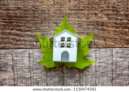 Miniature white toy model house with green maple leaf on wooden backdrop. Eco Village, abstract environmental background. Real estate mortgage property insurance dream home ecology concept