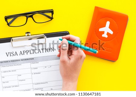 Hand fills visa application form. Form near glasses, pen, passport cover with airplane sign on yellow background top view copy space
