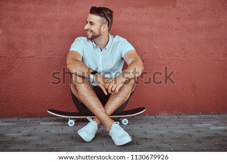 Smiling handsome fashionable guy dressed in a white shirt and shorts sitting on a skateboard under a bridge, looking away.