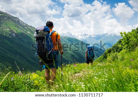 Tourists with hiking backpacks in mountain hike in Svaneti on summer day. Young guys go along the footpath through the mountains of Georgia. Tourists in beautiful mountain landscape. Royalty-Free Stock Photo #1130711717