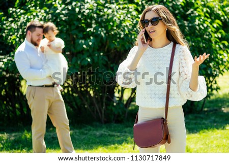 rich and beautiful woman cagy stylish sunglasses talking on the phone with her lover paramour on the phone in the park behind husband and daughter . the concept of divorce and adultery Royalty-Free Stock Photo #1130717849