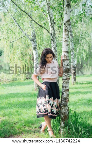 Young charming woman enjoying strolling while walking in park in sunny day, pretty brunette hair woman posing in park. Girl wearing in pink satin blouse, bell-skirt with embroidered ribbons flowers