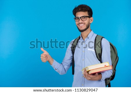 Cheerful young man wearing a classic outfit and his backpack, holding books in his hand, raising his thumb up and smiling, standing on a blue background
