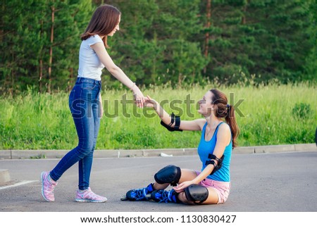 attractive young athletic slim brunette woman in pink shorts and blue top with protection elbow pads and knee pads on roller skates sitting on the asphalt with a girlfriend in the park .fall concept