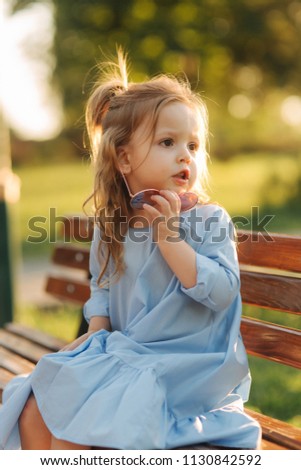 Little girl model in blue dress and sun glasses sits on a bench in the park