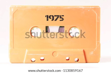 A vintage cassette tape from the 1980s era (obsolete music technology) with the text 1975 printed over it, stencil font. Color: cream, sand. White background.
