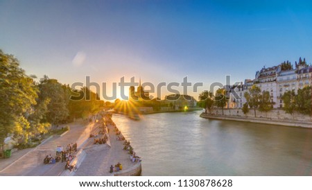 Rear view of Notre Dame De Paris cathedral at sunset with sun in the frame timelapse. View from Tournelle bridge. People sitting on waterfront. Orange sky in background. Paris, France, Europe.