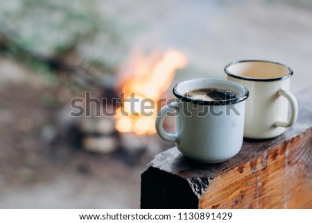 Two coffee sitting on an old log by an outdoor campfire. Selective focus on mug with blurred background. Camping.