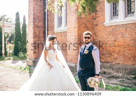 Beautiful newlyweds stand, embracing, near an old brick church with a large window. Portrait of a stylish groom and a young bride near an abandoned church.
