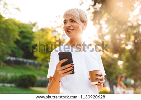 Photo of adult smiling woman with blond hair wearing casual clothing holding cell phone and takeaway coffee during walk in green park