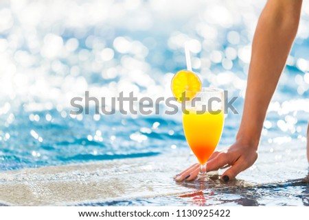 womans manicured hand touching a cocktail with an orange on a glass standing on the edge of the pool. free space for advertising text Royalty-Free Stock Photo #1130925422