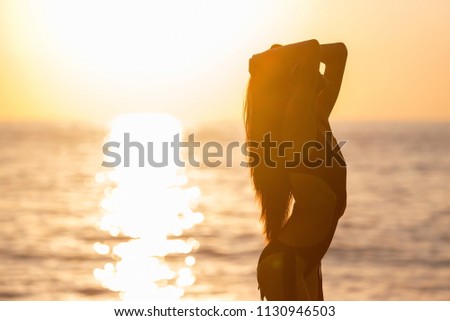 pretty young woman with long dark hair tanning in black swimwear against an ocean and sunset . concept of happy holiday and resort time. silhouette over sunrise Royalty-Free Stock Photo #1130946503