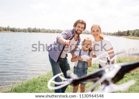 Gorgeous family is standing together and looking at drone that is on picture. Small girl is having control panel and smiling. Young man is pointing at drone.