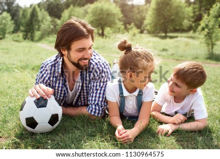 Father is lying on grass on meadow with children. He is holding ball with hand. Man is looking at his son with his daughter. Boy looks happy and funny.