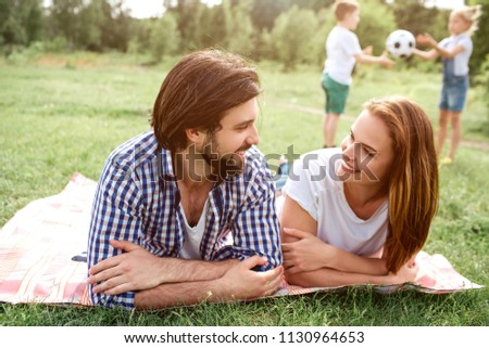 Happy parents are lying on blanket and looking at each other. They are smiling. There are their kids playing with ball down further.