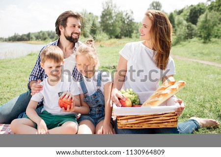 Parents are sitting on blanket with their kids. Woman is holding a basket with food. She is looking at man. He is smiling to her. Kids are holding one apple together.