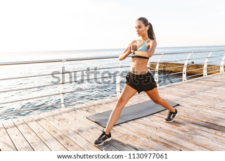 Image of energetic brunette sportswoman 20s in tracksuit doing sports and stretching body on boardwalk near seaside with bluetooth earbud
