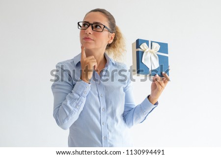 Pensive attractive woman holding gift and looking aside. Serious young lady in glasses thinking of present idea or what is inside of gift box. Contemplation concept