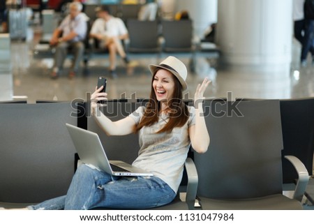 Laughing traveler tourist woman working on laptop, doing selfie on mobile phone, spreading hands, waiting in lobby hall at airport. Passenger traveling abroad on weekends getaway. Air flight concept