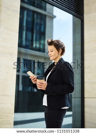 Side view of elegant businesswoman in black suit browsing phone outside and holding takeaway cup of coffee