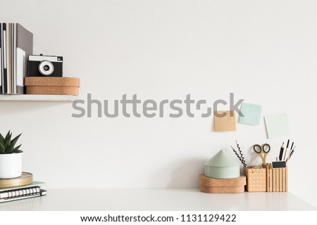 Stylish workspace desk with accessories, houseplant, sticky notes, camera on white wall background.