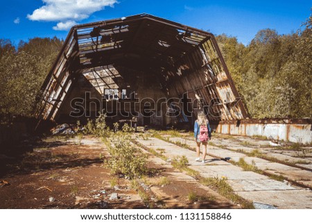 a girl in a denim jacket with a red backpack stands near a giant abandoned hangar and is going to take a picture of it on a mobile phone