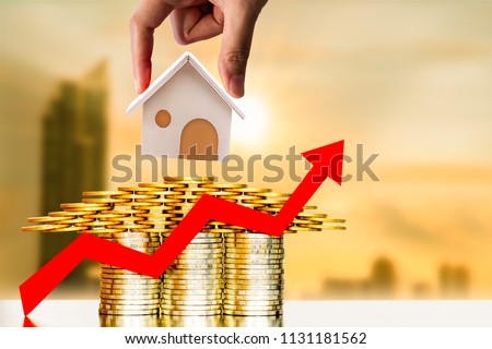 Investor hand hold a home model put on the stacked coin and red arrow graph with growing value on photo blur cityscape background, financial of real estate business investment and fund concept.
