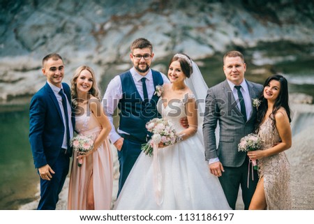 Wedding day bride and groom with bridesmaids and groomsmen posing in sunlight evening in mountains near river gorgeous wedding newlyweds couple with best friends.  People drink champagne and hugging.