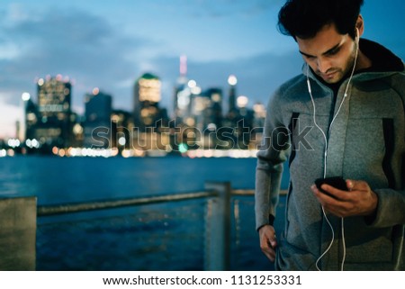 Young sportsman consulting with personal trainer about cardio exercises and listening audio message from him about guides for workout via earphones using sporty application on smartphone device