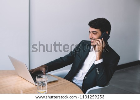 Positive smiling software developer calling to friend in break of updating webpage on laptop computer, cheerful indian boss spending time at company talking with colleague via smartphone