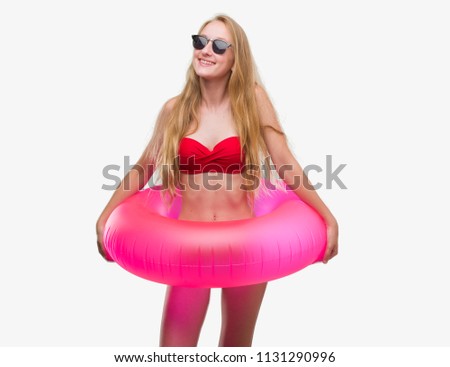 Blonde teenager woman wearing bikini and holding pink floater with a happy face standing and smiling with a confident smile showing teeth