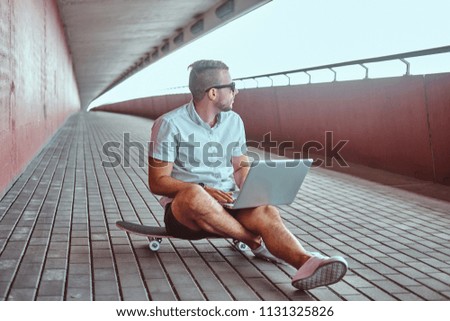 Portrait of a handsome fashionable freelancer in sunglasses dressed in a white shirt and shorts working on a laptop while sitting on a skateboard under the bridge.