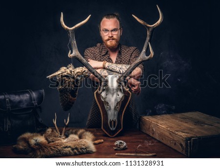 Redhead taxidermist hipster male in sunglasses dressed in a brown shirt, standing near a table with handmade trophy, owl scarecrow, and the fox skin. Isolated on the dark background. 
