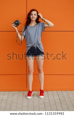 Hipster teen with vintage photo camera in one hand standing in front of orange wall, copy space, vintage toned image