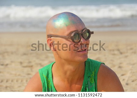 A bald and unusual young man, a freak, with a shiny bald head and round wooden glasses on the background of the beach and the sea. Humor and eccentricity. Unusual appearance. Humorist.