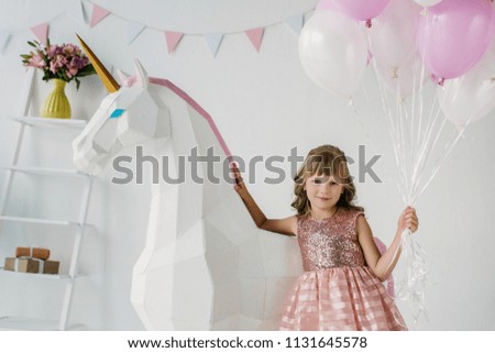 adorable little kid holding bunch of air balloons and standing with decorative unicorn 
