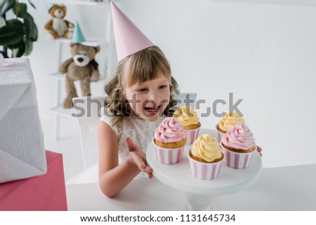 smiling birthday kid in cone holding stand with cupcakes at table 