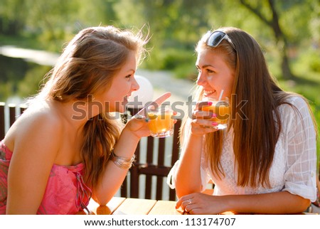 Two happy girl friends sitting in terrace cafe and chatting outdoors on a bright day of summer and green park on the background