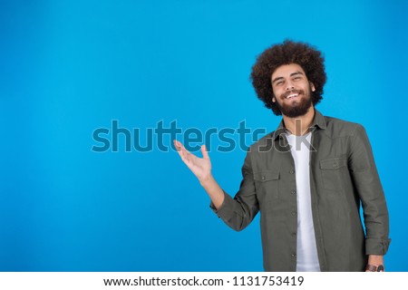 Young handsome man points with one hand inviting someone, on a blue background.