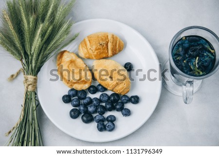 still-life tea from an orchid of blue color in a transparent mug. blueberries in a silver spoon. breakfast against white background. croissants in a white plate with blueberries. and green spikelets