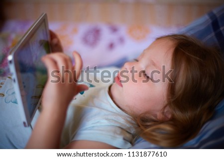 little Caucasian two-year-old girl lying on a plaid pillow and watching cartoons on a tablet computer. modern technology and childhood.